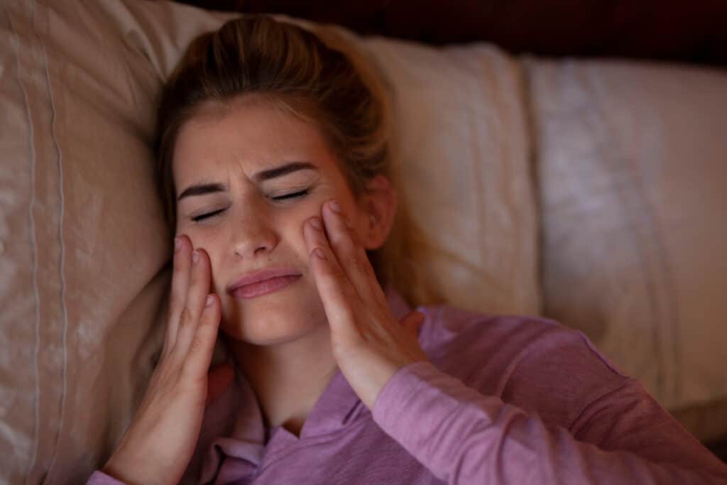 Woman laying in bed and rubbing her jaw in pain from bruxism, or teeth grinding